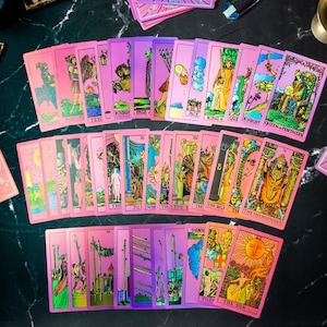 May include: A set of 22 tarot cards with pink and purple iridescent backgrounds. The cards feature traditional tarot imagery with gold accents. The cards are arranged in a fan shape, with the top card showing the Queen of Pentacles.