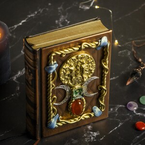 Op de afbeelding: Een gesloten, in leer gebonden boek met een gouden en bruine omslag. De omslag heeft een decoratief ontwerp met een drievoudig maansimbool, edelstenen en een gouden metalen accent.