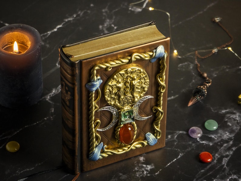 Op de afbeelding: Een gesloten, in leer gebonden boek met een gouden en bruine omslag. De omslag heeft een decoratief ontwerp met een drievoudig maansimbool, edelstenen en een gouden metalen accent.