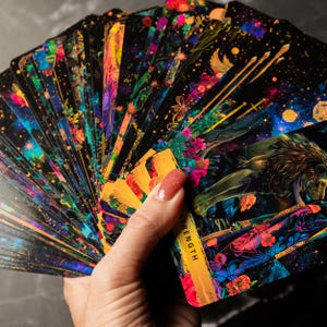 May include: A deck of tarot cards fanned out, featuring vibrant illustrations with a celestial and floral theme. The cards have a black background with colorful artwork, including a lion and the word "STRENGTH".