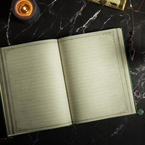 Op de afbeelding: Een open leeg dagboek met een groene kaft en gelinieerde pagina's. Het dagboek ligt op een zwart marmeren oppervlak met een brandende kaars, een klein kristal en andere decoratieve items.