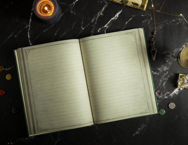 Op de afbeelding: Een open leeg dagboek met een groene kaft en gelinieerde pagina's. Het dagboek ligt op een zwart marmeren oppervlak met een brandende kaars, een klein kristal en andere decoratieve items.