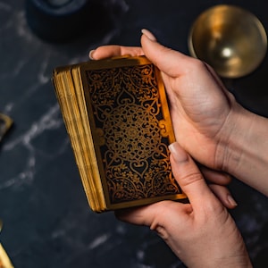 May include: A deck of tarot cards with gold edges and a black and gold ornate design is held in both hands. The cards are on a dark, textured surface, with a gold bowl and other cards visible in the background.