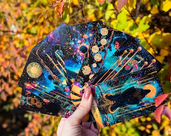 May include: A hand holding a deck of tarot cards with vibrant, celestial artwork. The cards feature illustrations with blues, golds, and purples, depicting mystical scenes. The cards are fanned out, showcasing the detailed designs and imagery.