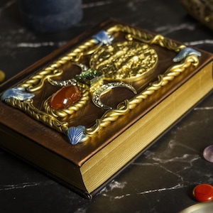 Op de afbeelding: Een bruin lederen gebonden boek met gouden en zilveren versieringen. De omslag heeft een decoratief ontwerp met een groot, rond, gouden middelpunt en een rode edelsteen.