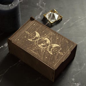 May include: A wooden box with a lid, featuring an engraved design of three moons and a star. The box is dark brown and has a natural wood grain.