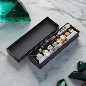 May include: A black box with a set of seven chakra stones, a crystal wand, and a beaded bracelet. The box is open and the stones are arranged in a row. The stones are different colors, representing the seven chakras. The wand is made of clear quartz and the bracelet is made of brown leather with beads of various colors.