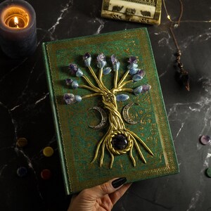 Op de afbeelding: Een groen lederen gebonden dagboek met een gouden levensboomontwerp. De boom is versierd met paarse en blauwe edelstenen. Het dagboek is gesloten en wordt in een hand gehouden.