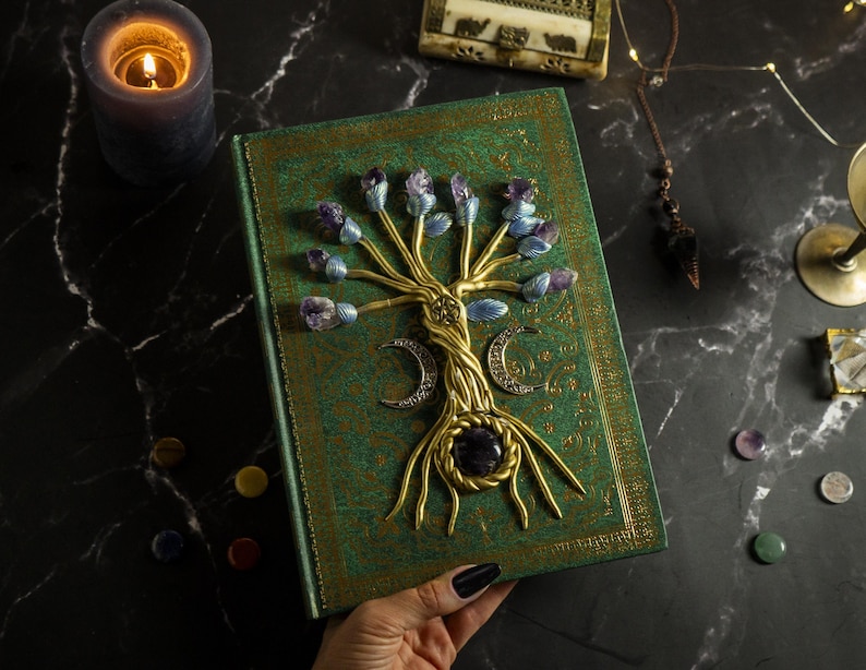 Op de afbeelding: Een groen lederen gebonden dagboek met een gouden levensboomontwerp. De boom is versierd met paarse en blauwe edelstenen. Het dagboek is gesloten en wordt in een hand gehouden.