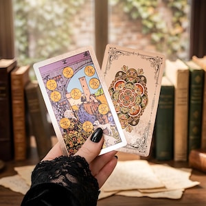 May include: Two rectangular tarot cards held up, one displaying the Ten of Pentacles with ten gold coins and figures in an archway, the other showing a detailed, symmetrical, multi-colored floral mandala pattern with ornate black border detailing.