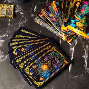 May include: A tarot card deck with a celestial theme. The cards have a dark blue backdrop with a sunburst, flowers, and stars, and gold edges. One card reads "2 of Pentacles".