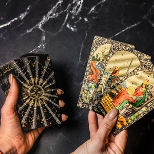 May include: A deck of tarot cards with a black and gold sunburst design on the back. Two cards are displayed, featuring illustrations of figures in ornate frames. The cards are held against a dark, marbled surface.