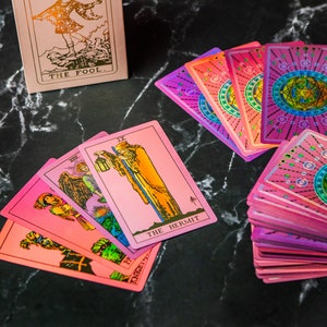 May include: A spread of tarot cards with pink and purple holographic foil accents. The cards include The Fool, The Hermit, and several other cards with roman numeral markings. The cards are laid out on a black marble surface.