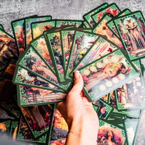 May include: A fan of rectangular tarot cards with dark green borders and intricate floral illustrations. The cards feature colorful scenes of figures in nature, architectural elements, and symbolic imagery, with some cards displaying Roman numerals.