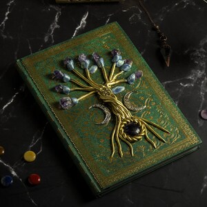 Op de afbeelding: Een groen lederen gebonden dagboek met een gouden boomontwerp op de omslag. De boom is versierd met amethist en blauwe kristallen en heeft een gouden halve maan aan de basis.