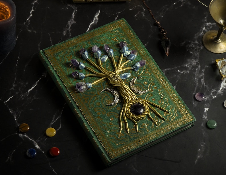 Op de afbeelding: Een groen lederen gebonden dagboek met een gouden boomontwerp op de omslag. De boom is versierd met amethist en blauwe kristallen en heeft een gouden halve maan aan de basis.