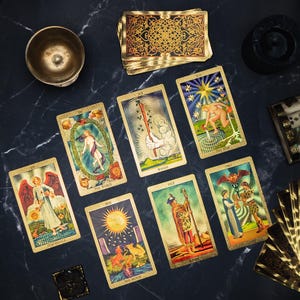 May include: A collection of tarot cards arranged on a dark surface, including The World, Ace of Wands, The Star, Temperance, The Moon, King of Wands, and Two of Cups. A brass bowl, a small box, and a deck of cards with gold-patterned backs are present.