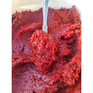 May include: Close-up of a vibrant red, thick paste, possibly tomato paste or a similar condiment. A silver spoon is partially inserted, holding a portion of the paste. The texture appears chunky and rich, with a deep red color.