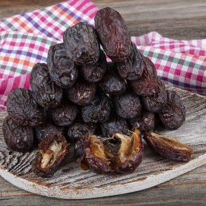 May include: A large pile of dark brown, wrinkled Medjool dates stacked on a round, weathered wooden board. Several dates are split open, revealing their fibrous, light-colored interior flesh and a single, hard, elongated seed inside each fruit.