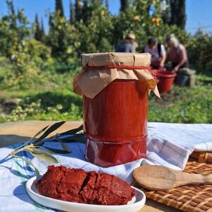 Pode incluir: Um frasco de pasta de tomate vermelha, com tampa de papel castanho e barbante, está sobre uma mesa. Um prato pequeno de pasta e uma colher de pau estão ao lado. O fundo mostra uma cena ensolarada ao ar livre.