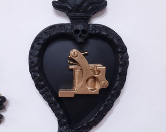 Pode incluir: Decoração de parede em forma de coração preto e dourado com uma máquina de tatuagem dentro. O coração é coroado com uma chama e uma caveira. O coração está pendurado em uma corrente dourada com uma caveira na parte inferior.