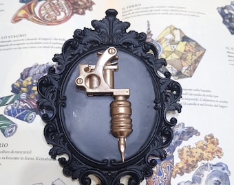 May include: A gold-colored tattoo machine is displayed in a black ornate frame. The frame is set against a page from a book about elements.