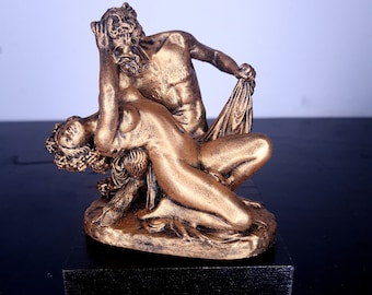 May include: A gold-toned sculpture depicting a couple embracing. The male figure is holding a drape in his hand, while the female figure is draped in fabric. The sculpture is set on a black base.
