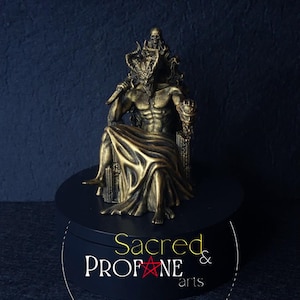 Könnte beinhalten: Goldfarbene Statue einer gehörnten Figur auf einem Thron, die eine Klinge hält. Die Statue weist komplizierte Details auf und steht auf einer schwarzen Plattform. Der Text "Sacred & Profane arts" ist sichtbar.