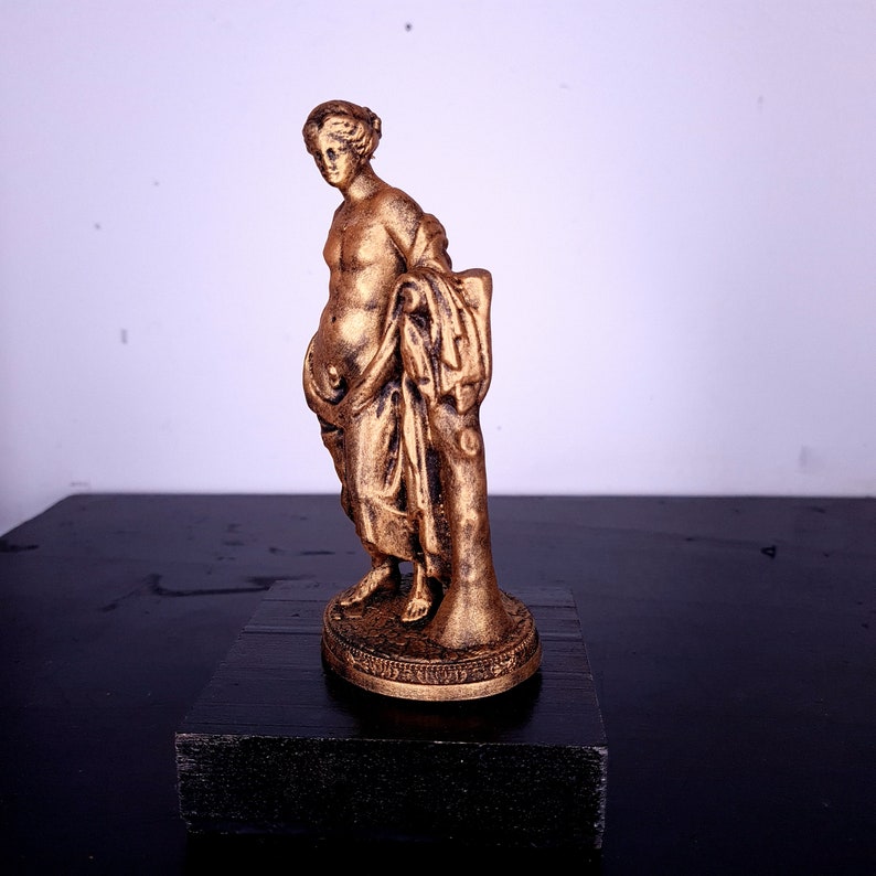 May include: A gold-colored statue of a woman standing on a pedestal. The woman is wearing a flowing robe and has her hair pulled back. The statue is on a black surface.