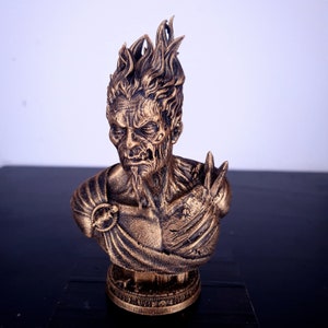 May include: A gold-colored bust sculpture of a demonic figure with a fiery head and a detailed, textured surface. The bust is on a round base.