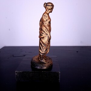 May include: A gold-colored statue of a woman standing on a black base. The statue is wearing a long, flowing robe and has her hair pulled back in a bun.