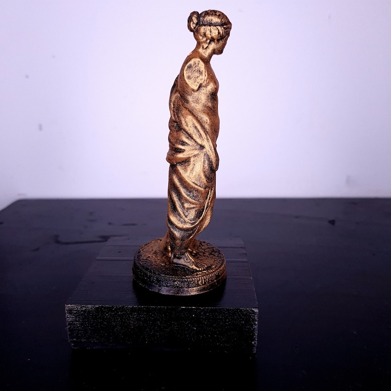 May include: A gold-colored statue of a woman standing on a black base. The statue is wearing a long, flowing robe and has her hair pulled back in a bun.