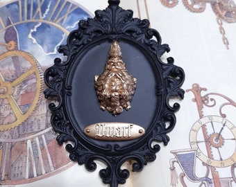 Pode incluir: Uma moldura preta adornada com uma figura de gnomo dourada no seu interior. A moldura tem uma placa dourada com a palavra "Anão" escrita nela.