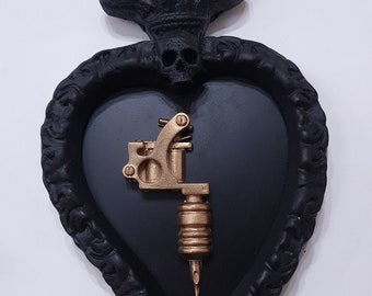 Pode incluir: Decoração de parede em forma de coração preto com uma máquina de tatuagem dourada no interior. O coração tem uma caveira e chamas no topo.