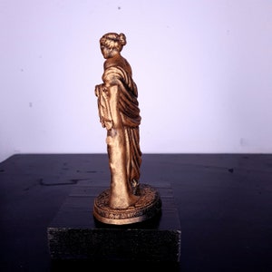 May include: A gold-colored statue of a woman standing on a round base. The statue is detailed and has a draped garment.