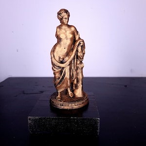 May include: A gold-colored statue of a woman standing on a black base. The woman is wearing a flowing robe and has her hair pulled back.