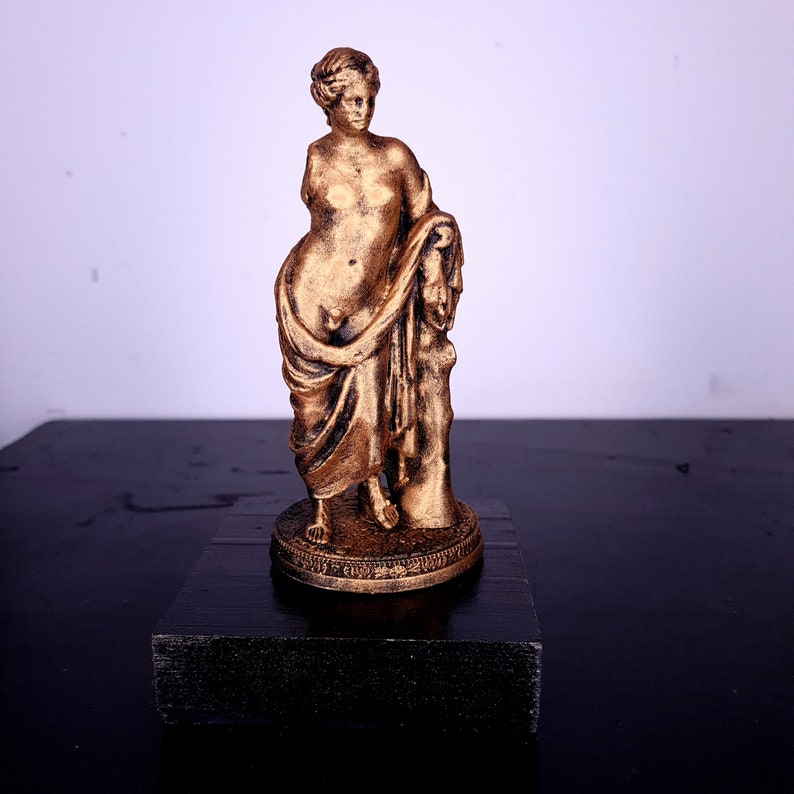 May include: A gold-colored statue of a woman standing on a black base. The woman is wearing a flowing robe and has her hair pulled back.