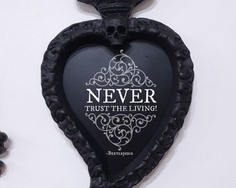 Pode incluir: Moldura preta em forma de coração com um crânio e chamas no topo. A moldura tem uma inscrição branca que diz "NEVER TRUST THE LIVING!" e "BEETLEJUICE" na parte inferior.