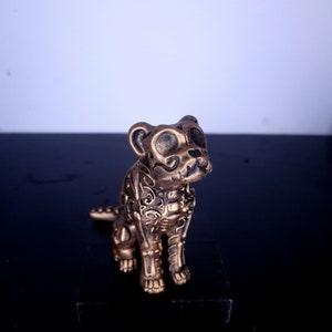 May include: A bronze-colored dog figurine with intricate floral and swirl patterns. The dog is in a sitting position, with its head tilted slightly to the side. The figurine has large, stylized eyes and a detailed snout. The overall design has a Day of the Dead aesthetic.