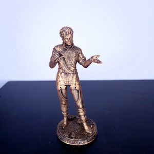 Può includere: Una statuetta dorata di una persona in piedi su una base circolare. La persona indossa un abito dettagliato con una spada al fianco e tiene un pugnale nella mano sinistra. La statuetta è realizzata in plastica rigida.