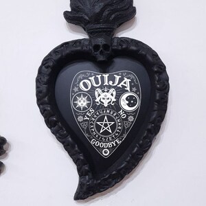 May include: Black heart-shaped frame with a skull and flames on top. The frame contains a black and white Ouija board with the words "Ouija", "Yes", "No", and "Goodbye".
