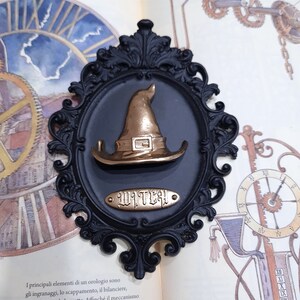 May include: A black ornate frame with a gold witch hat and the word "Witches" written on a gold plaque.