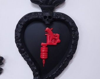 Pode incluir: Decoração de parede em forma de coração preto e vermelho com um design de chama no topo e uma máquina de tatuagem dentro. A decoração está pendurada em uma corrente dourada com uma caveira na parte inferior.
