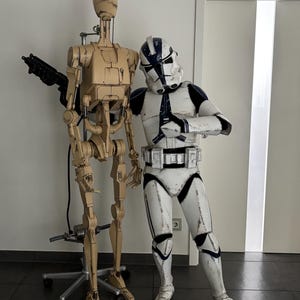 May include: A beige and brown robot with a gun arm stands next to a white and blue stormtrooper action figure. The stormtrooper is holding its arms in front of its chest.