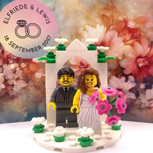 Puede incluir: Un adorno de pastel de boda de Lego con una novia y un novio de pie bajo un arco blanco con flores. El adorno de pastel está sobre una base blanca con flores verdes y blancas. La tarjeta detrás del adorno de pastel tiene una pegatina que dice "Elfriede & Lewis 18 de septiembre de 2021".