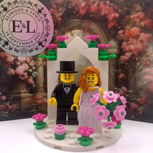 May include: A Lego wedding cake topper featuring a bride and groom in formal attire. The bride is wearing a white dress with pink flowers and the groom is wearing a black tuxedo. The cake topper is decorated with pink flowers and a white archway. The cake topper is on a white base with the text "E&L 18 September 2021" on the side.