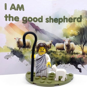 May include: Lego figurine of a shepherd with a staff and a lamb, standing on a green base. The figurine is in front of a watercolor painting of a landscape with sheep and a tree. The text "I AM the good shepherd" is written on the card behind the figurine.