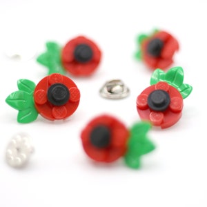 May include: Five red poppy pins with green leaves and black centres. The pins are made from Lego bricks and are attached to metal pin backs.