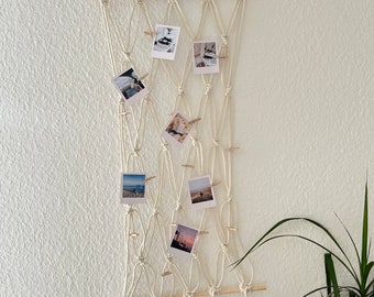Könnte beinhalten: Ein weißer Makramee-Wandbehang mit einer Holzleiste und Quasten. Der Behang hat ein Gittermuster und ist mit Fotos verziert, die mit Holz-Wäscheklammern befestigt sind.