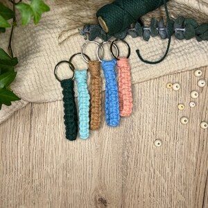 May include: Five colourful macrame keychains with silver key rings. The keychains are made with different coloured cords and are hanging on a wooden surface.